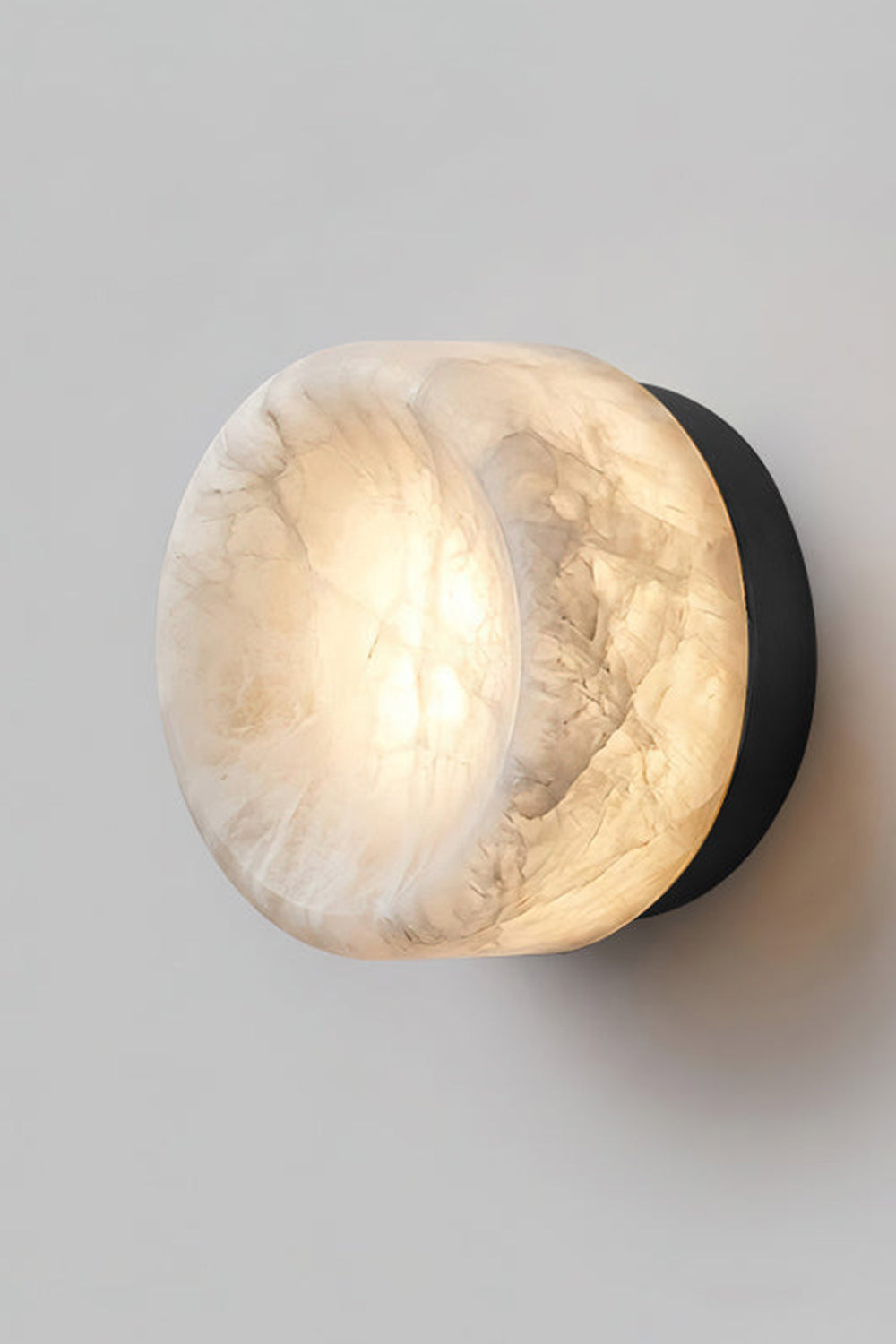 Isolde Concave Alabaster Disc Wall Sconce | Natural Alabaster and Black Metal | Studio close-up shot of the lit sconce featuring a matte black finish base on a light wall | Samu Lamp