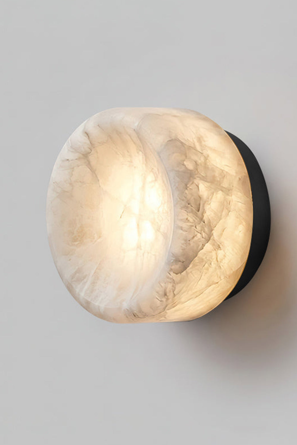 Isolde Concave Alabaster Disc Wall Sconce | Natural Alabaster and Black Metal | Studio close-up shot of the lit sconce featuring a matte black finish base on a light wall | Samu Lamp