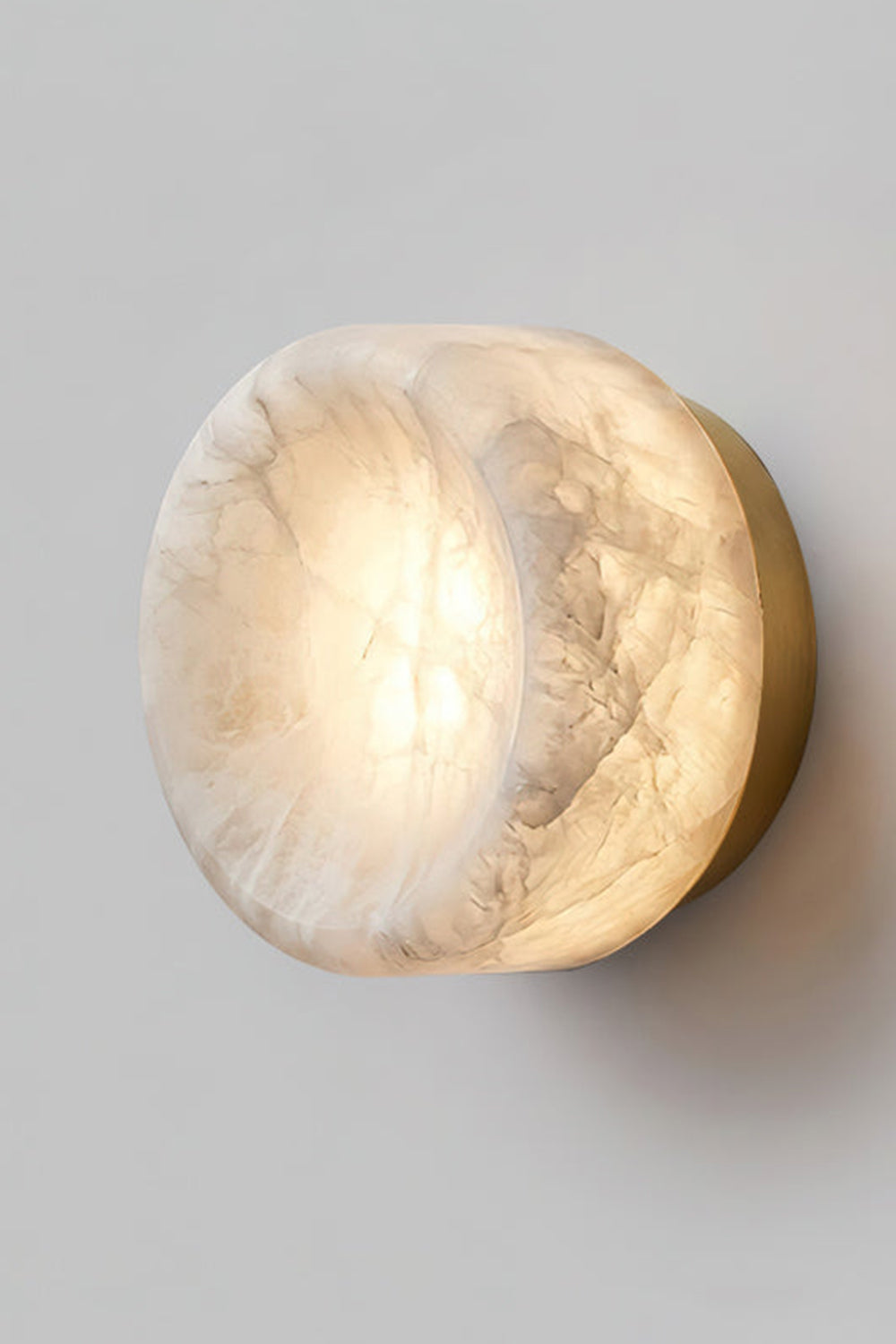 Isolde Concave Alabaster Disc Wall Sconce | Natural Alabaster and Black Metal | Studio close-up shot of the lit sconce featuring a matte black finish base on a light wall | Samu Lamp