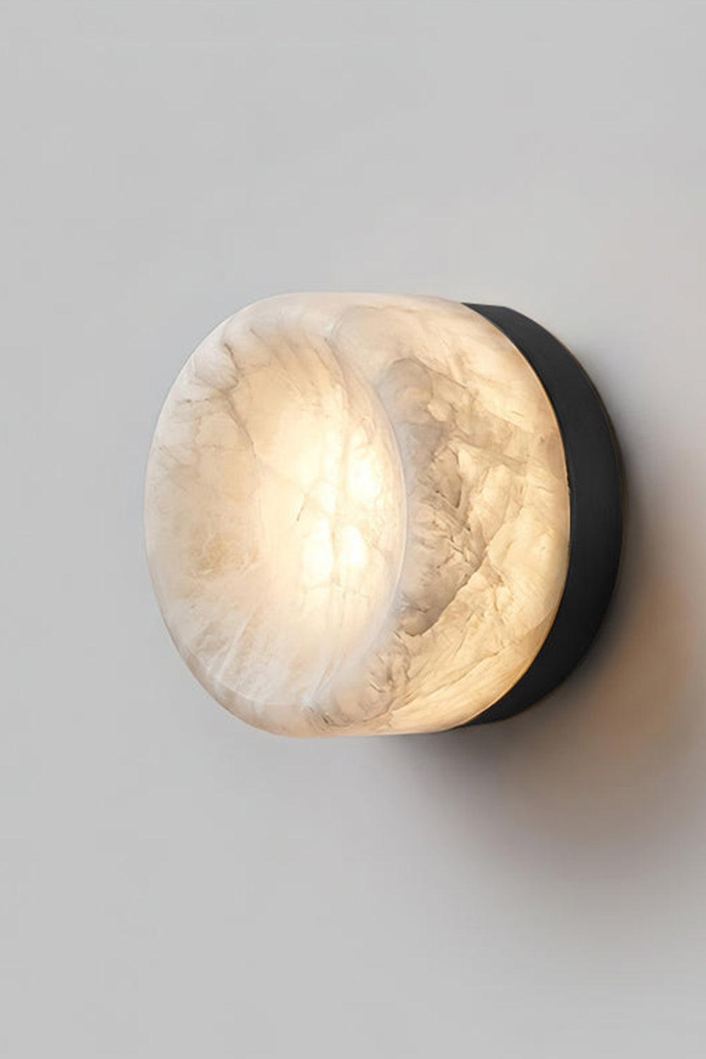 Isolde Concave Alabaster Disc Wall Sconce | Natural Alabaster and Black Metal | Studio close-up shot of the lit sconce featuring a matte black finish base on a light wall | Samu Lamp