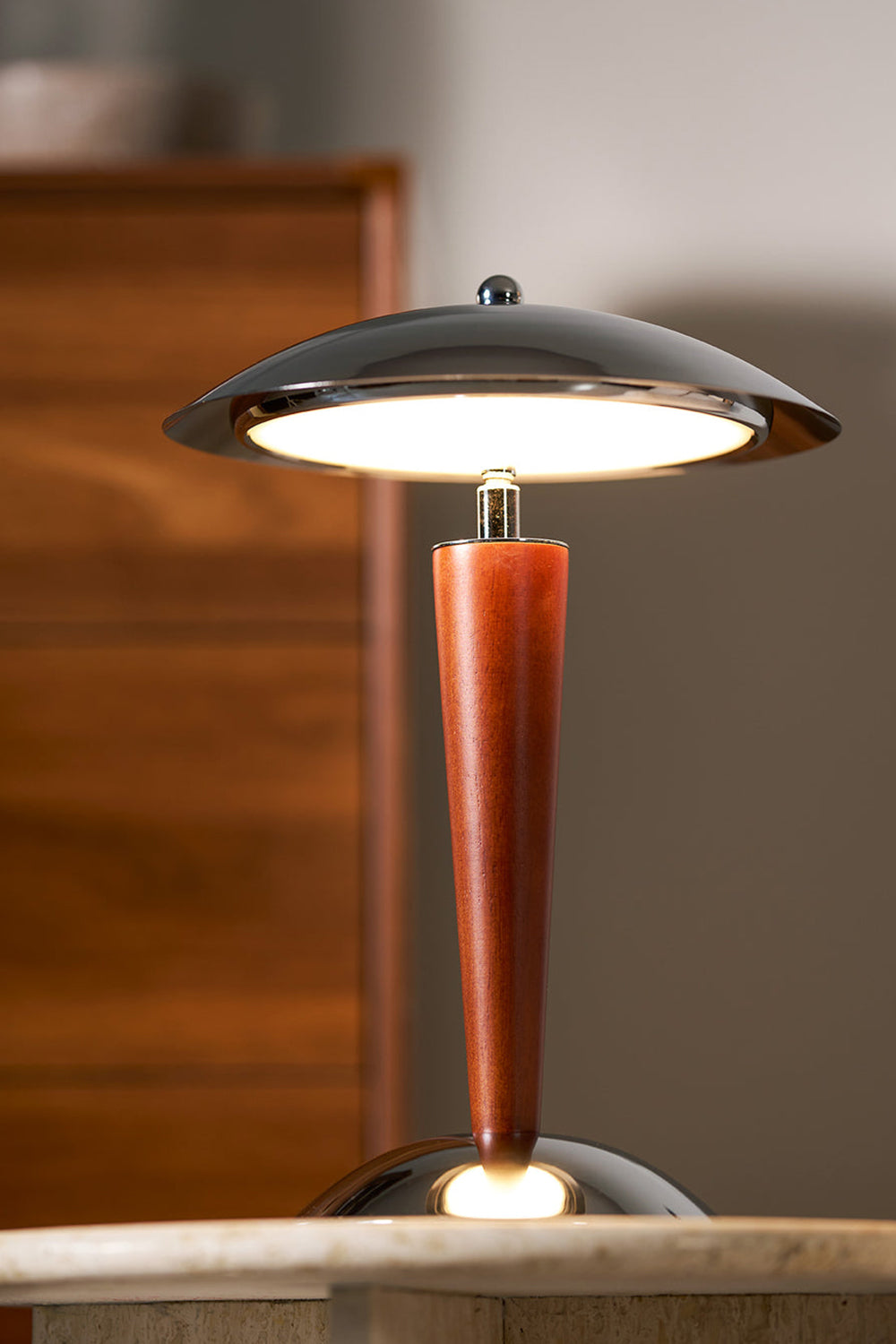Isolde Domed Walnut Table Lamp | Cozy Accent Glow | Staged view of the fully illuminated mushroom lamp casting a warm yellow halo against a dark blurred wooden chest background | Samu Lamp