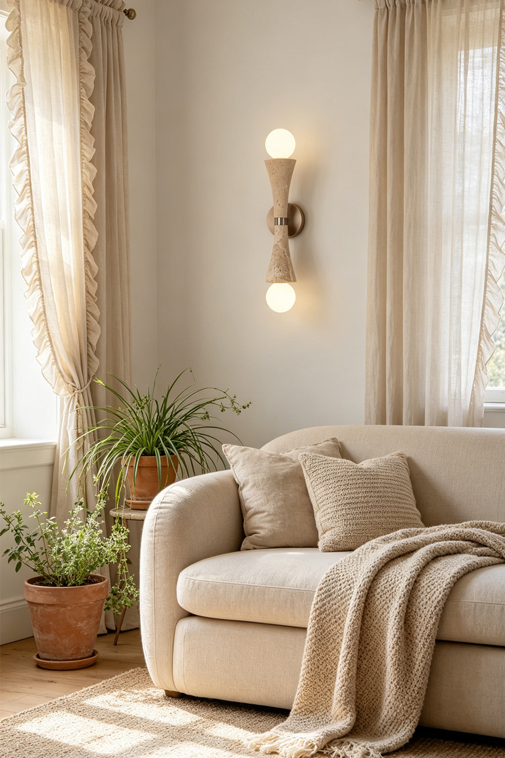 Katia Travertine Hourglass Wall Lamp | Travertine, Metal | Positioned gracefully on a living room wall, radiating soft accent lighting next to a plush beige fabric sofa with throw blankets | Samu Lamp