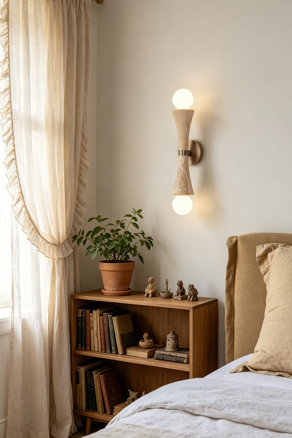 Katia Travertine Hourglass Wall Lamp | Travertine, Metal | Providing a warm ambient light in a cozy bedroom corner next to a wooden storage shelving unit and linen drapery window | Samu Lamp