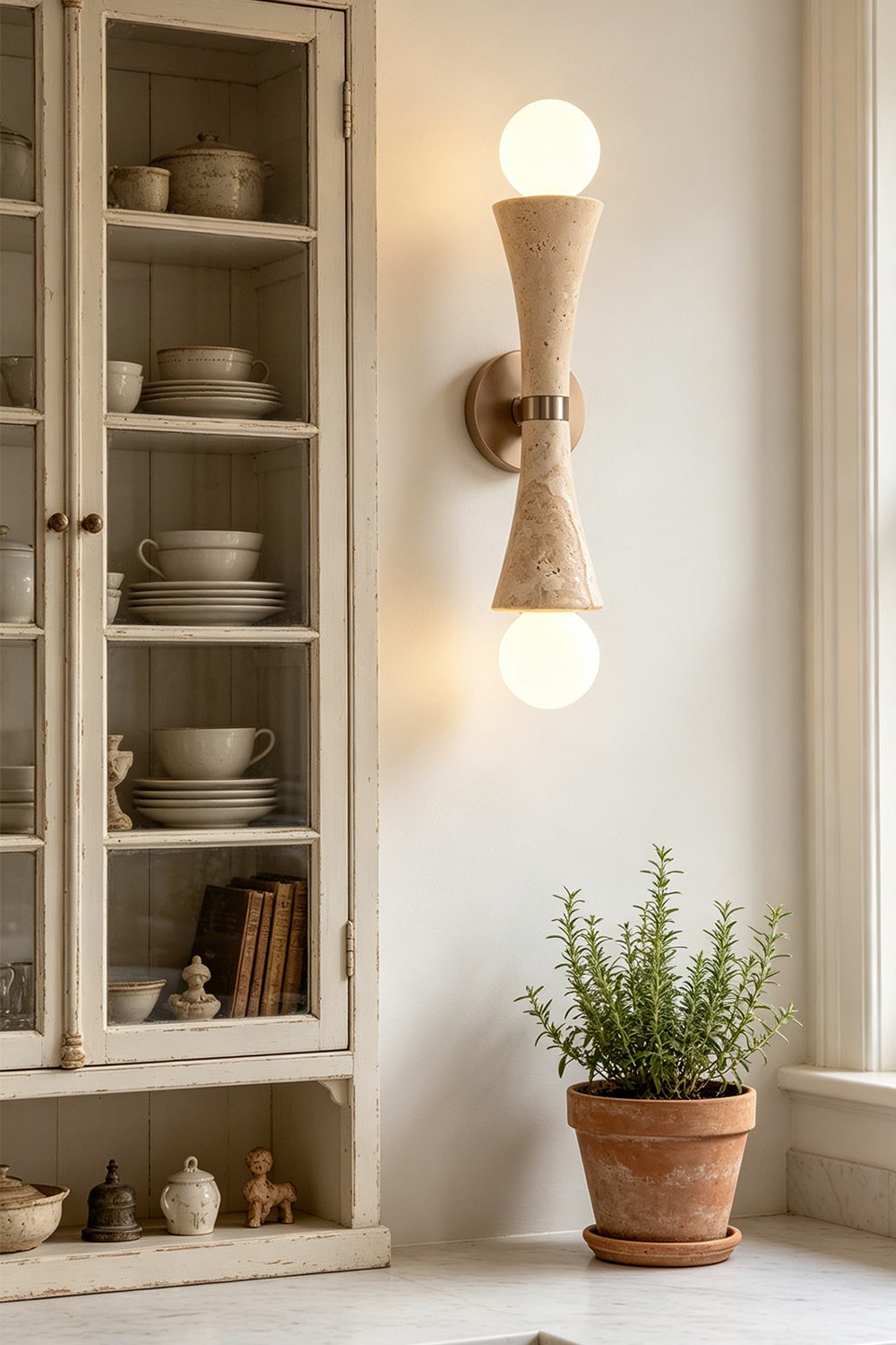 Katia Travertine Hourglass Wall Lamp | Travertine, Metal | Styled in a contemporary interior next to a vintage glass hutch cabinet filled with ceramic dishes and a terracotta plant pot | Samu Lamp