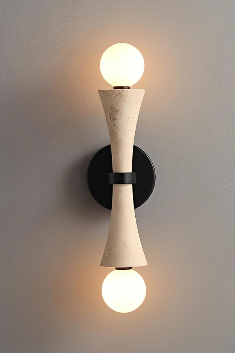 Katia Travertine Hourglass Wall Lamp | Travertine, Metal | Studio front view of the fully illuminated hourglass stone wall fixture casting a brilliant dual-directional warm light halo against a grey backdrop | Samu Lamp