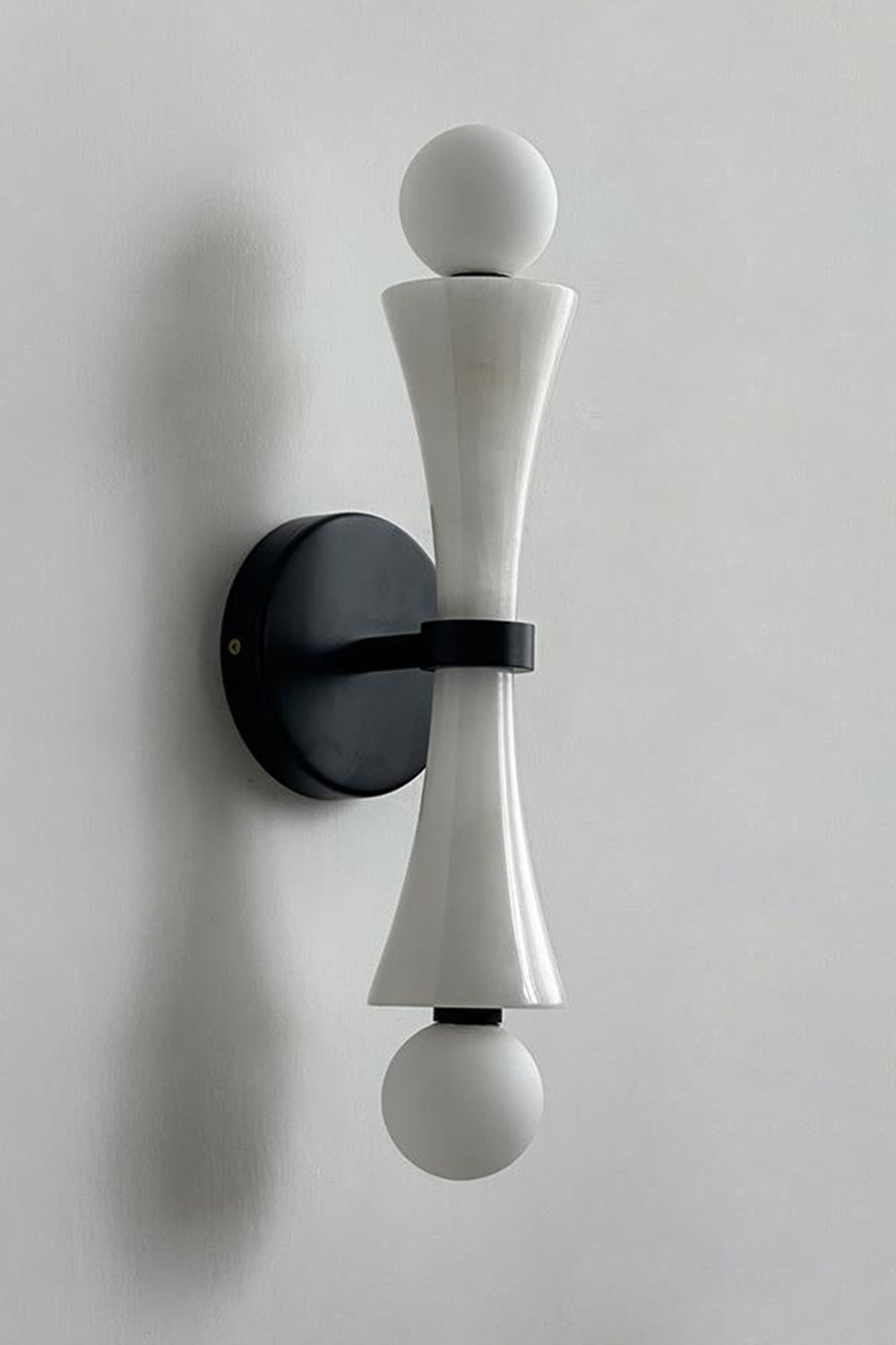 Katia Travertine Hourglass Wall Lamp | Travertine, Metal | Studio variant product view featuring a smooth white marble or stone hourglass body paired with black hardware fittings | Samu Lamp