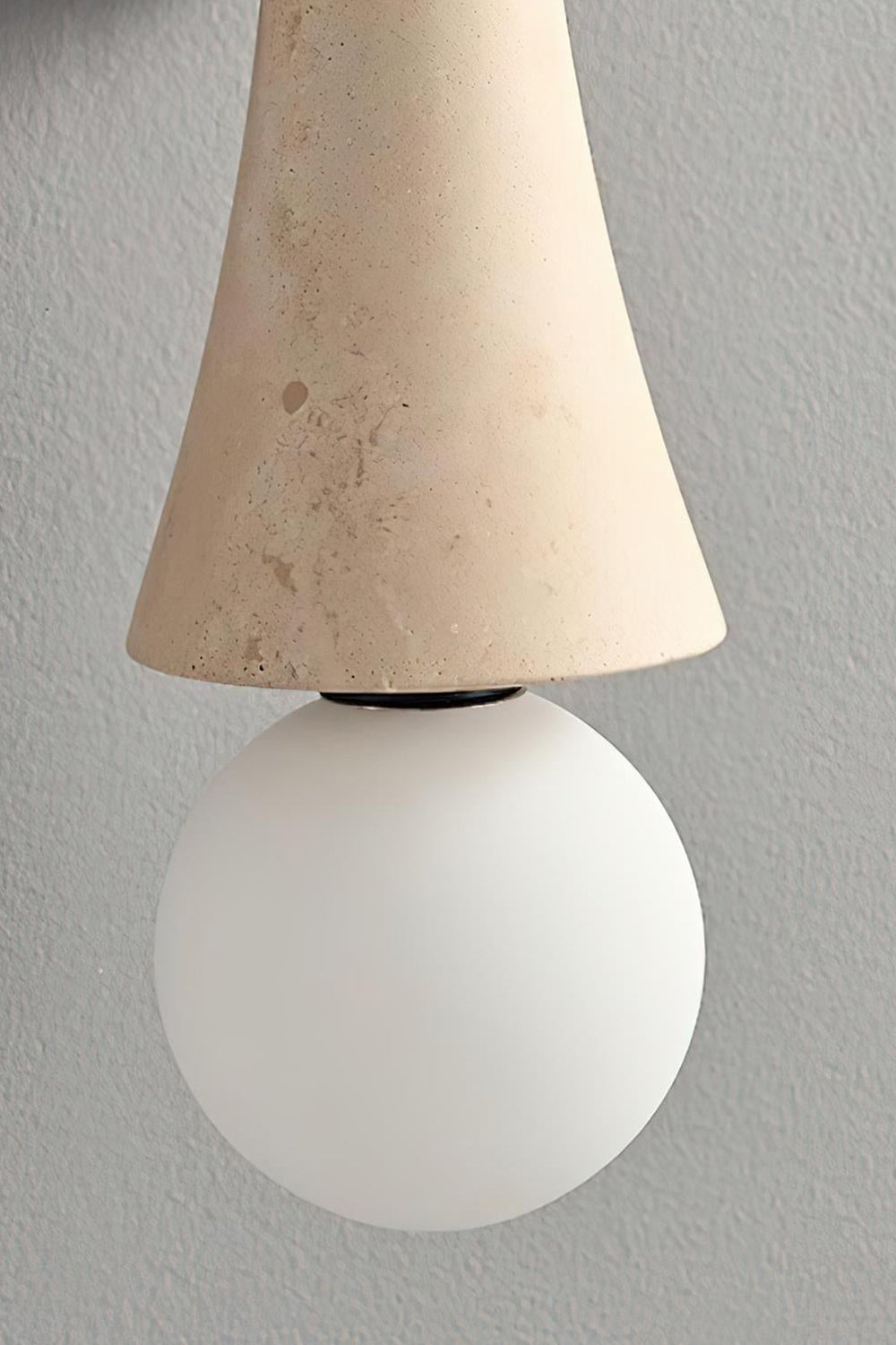 Katia Travertine Hourglass Wall Lamp | Travertine, Metal | Close-up focus on the white frosted glass globe bulb seamlessly meeting the tapered edge of the natural porous travertine stone shade | Detail shot | Samu Lamp