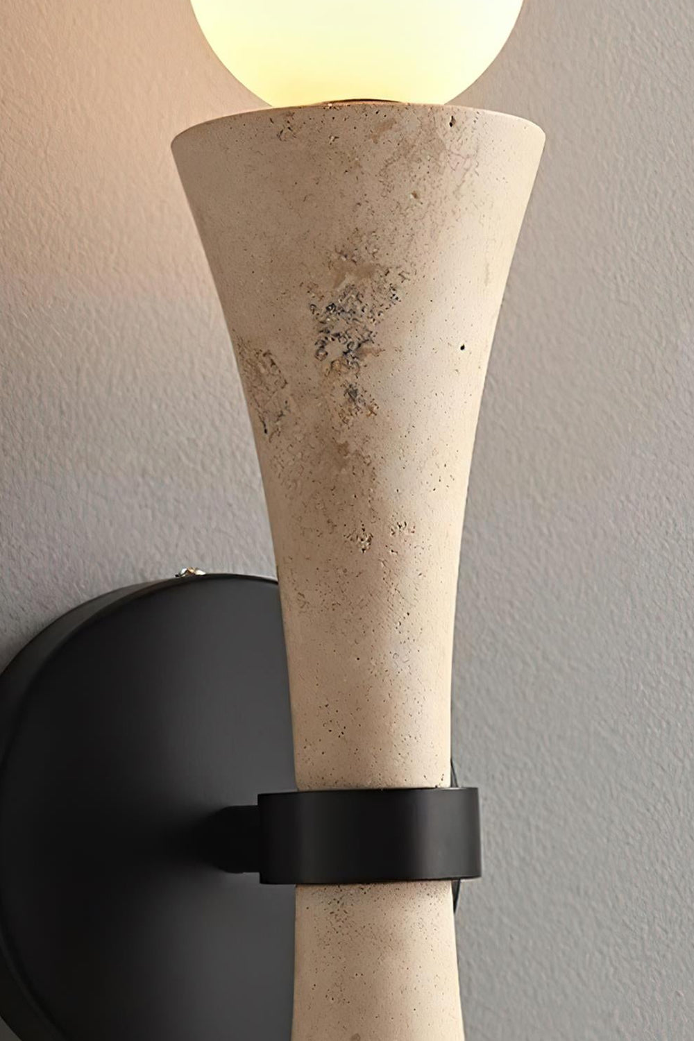 Katia Travertine Hourglass Wall Lamp | Travertine, Metal | Side profile up-close view showing the textured beige stone hourglass body held firmly by a matte black metallic ring clamp collar | Detail shot | Samu Lamp