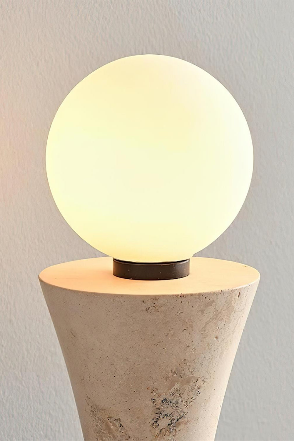 Katia Travertine Hourglass Wall Lamp | Travertine, Metal | Detailed shot of the top spherical globe light bulb illuminated, radiating a soft warm white glow over the stone texture | Detail shot | Samu Lamp