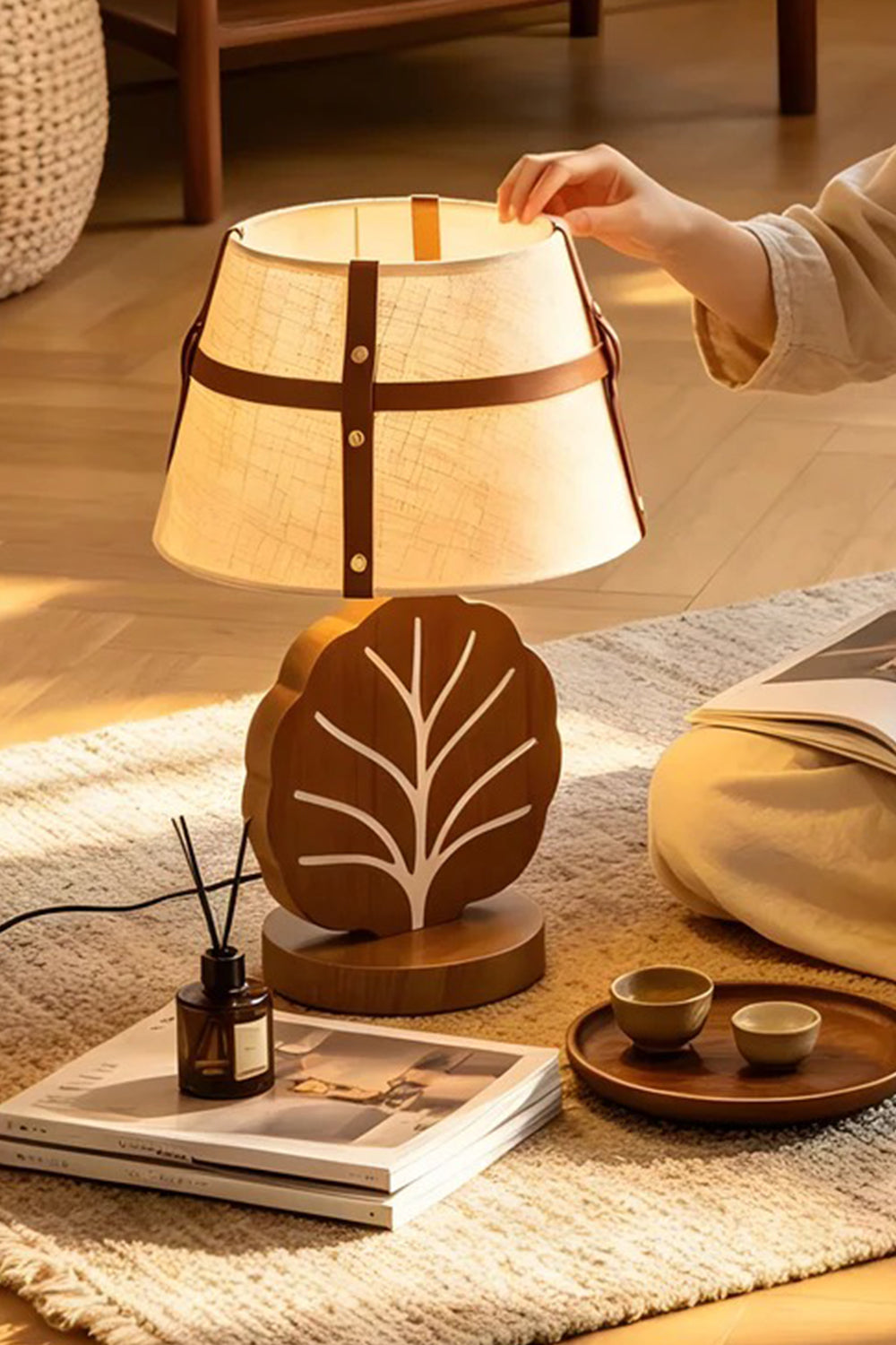 Leafora Botanical Leaf Table Light | Wood, Fabric, Leather | Cozy indoor scene on a textured floor rug next to books, showing a hand interacting with the fully illuminated leaf table lamp | Samu Lamp