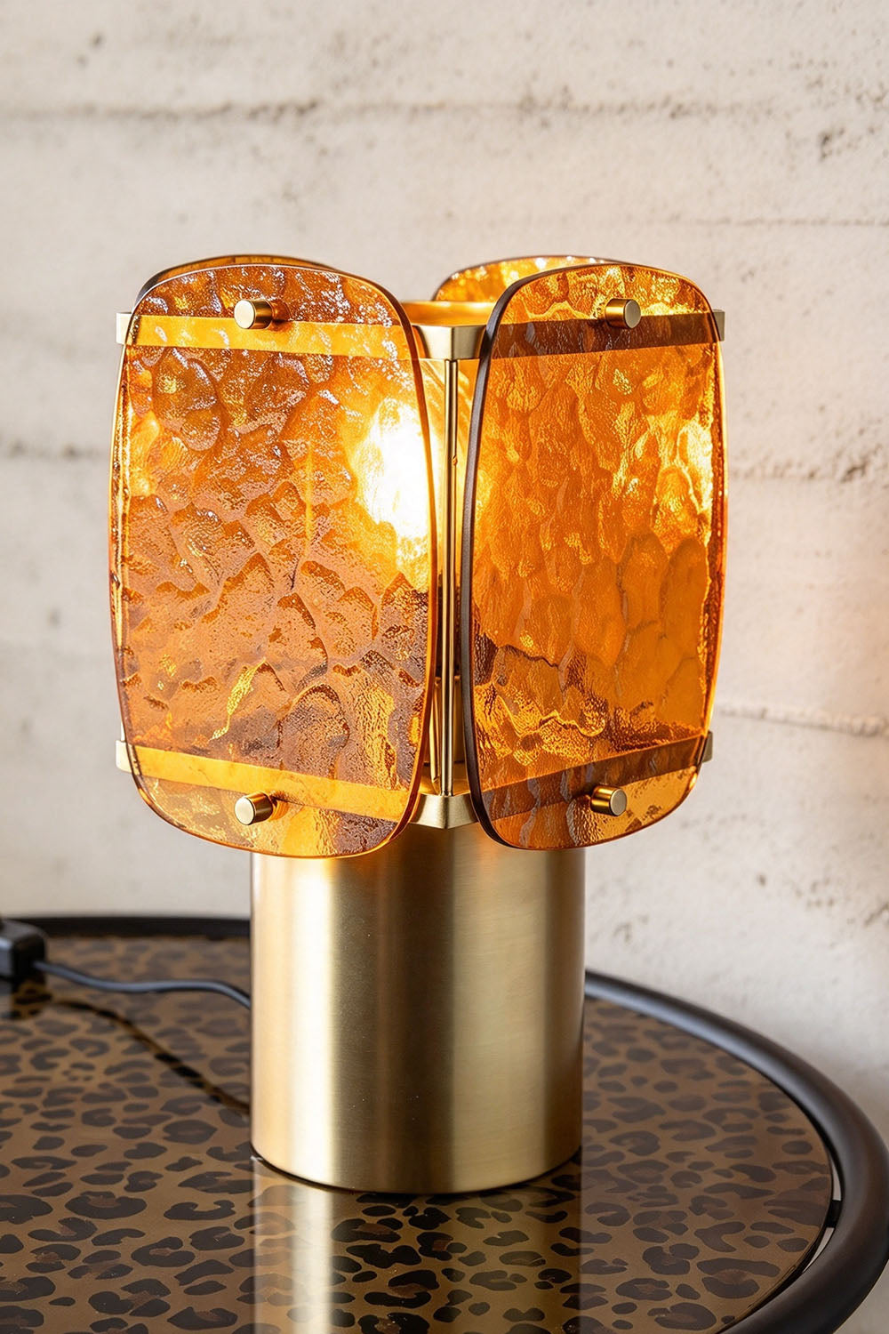 Lesquin Hammered Amber Glass Table Lamp | Hammered Amber Glass and Brass | Glowing table lamp displayed on a round side table against a white brick wall | Samu Lamp