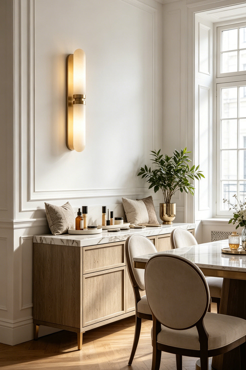 Linea Alabaster Capsule Wall Sconce | Alabaster, Brass | Radiating warm ambient lighting on a white wall near a dining area with a classic sideboard and chairs | Samu Lamp
