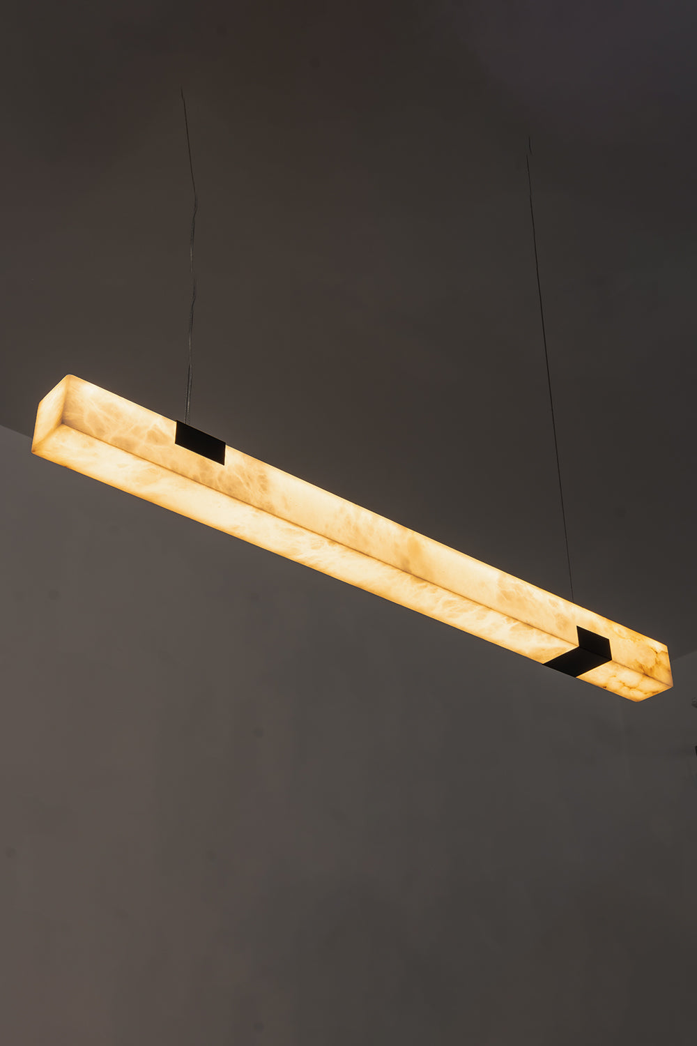 Liora Linear Alabaster Pendant Light | Natural Alabaster and Black Metal | High-angle view of the illuminated linear stone fixture glowing against a dark background | Samu Lamp