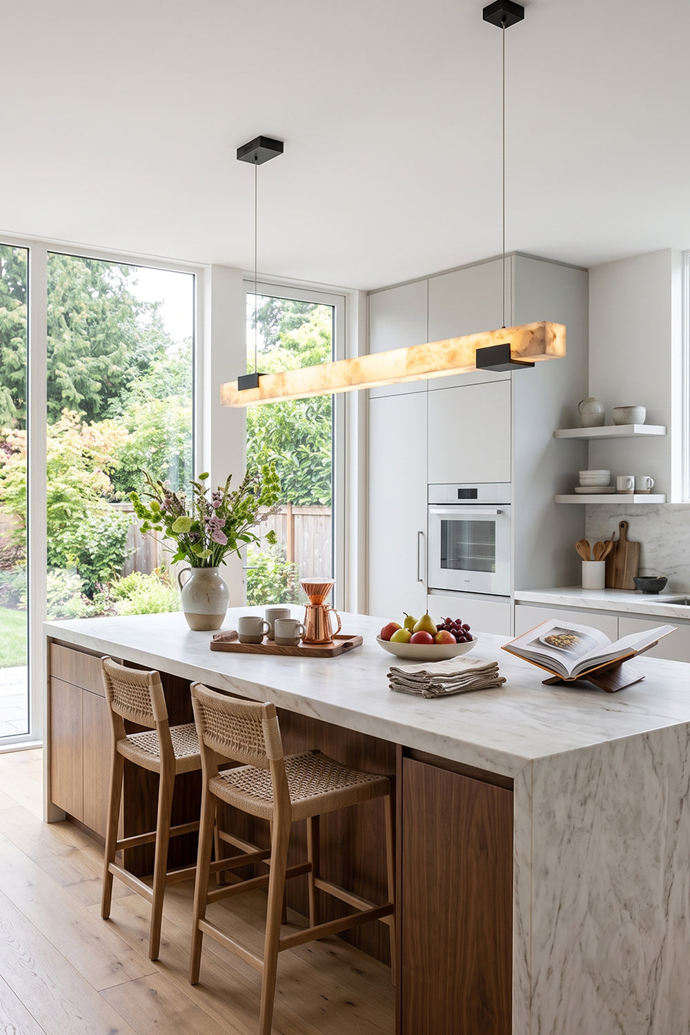 Liora Linear Alabaster Pendant Light | Natural Alabaster and Black Metal | Positioned over a long marble kitchen counter surrounded by matching wooden bar stools | Samu Lamp