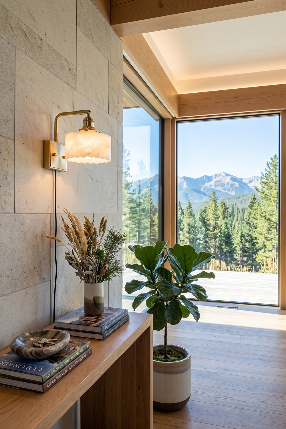 Liora Scalloped Alabaster Wall Sconce | Natural Alabaster and Brushed Brass | Styled in a modern hallway above a wooden console table near a large window with scenic mountain views | Samu Lamp
