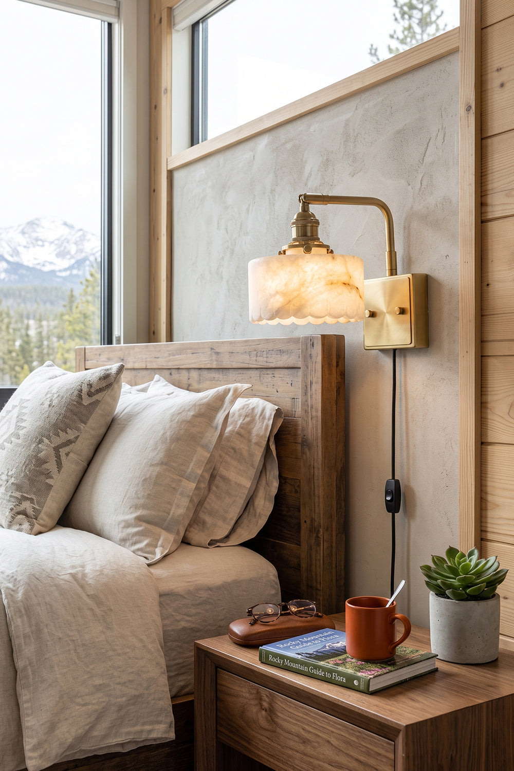 Liora Scalloped Alabaster Wall Sconce | Natural Alabaster and Brushed Brass | Mounted on a wooden bedroom wall beside a bed and a rustic nightstand with a snowy mountain window view | Samu Lamp