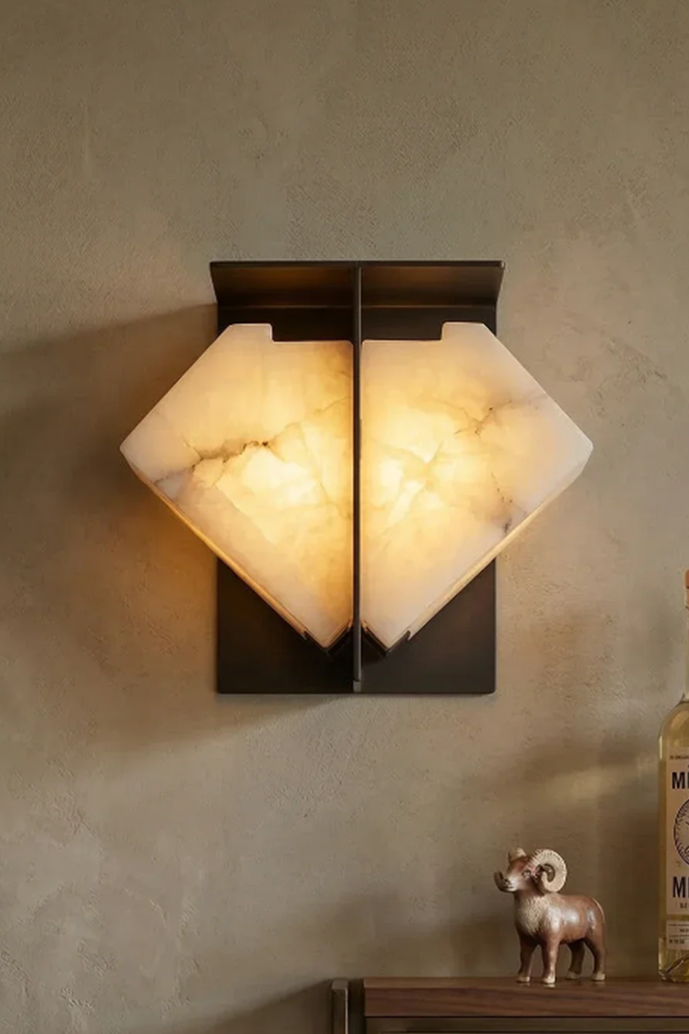 Lysandra Alabaster Folded Halo Wall Lamp | Cozy Mood Lighting | Fully lit geometric stone wall light casting an inviting yellow halo on a textured plaster wall above a shelf decoration | Samu Lamp