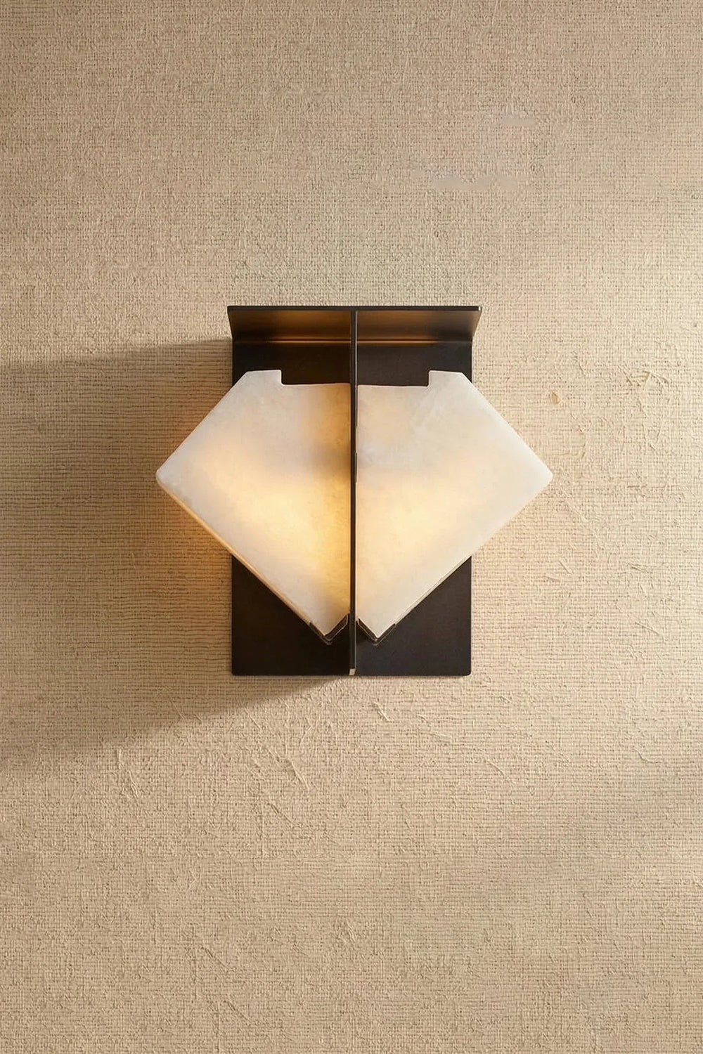 Lysandra Alabaster Folded Halo Wall Lamp | Studio Front View | Frontal view of the illuminated folded envelope-shaped alabaster sconce casting a warm halo on a plain beige backdrop | Samu Lamp