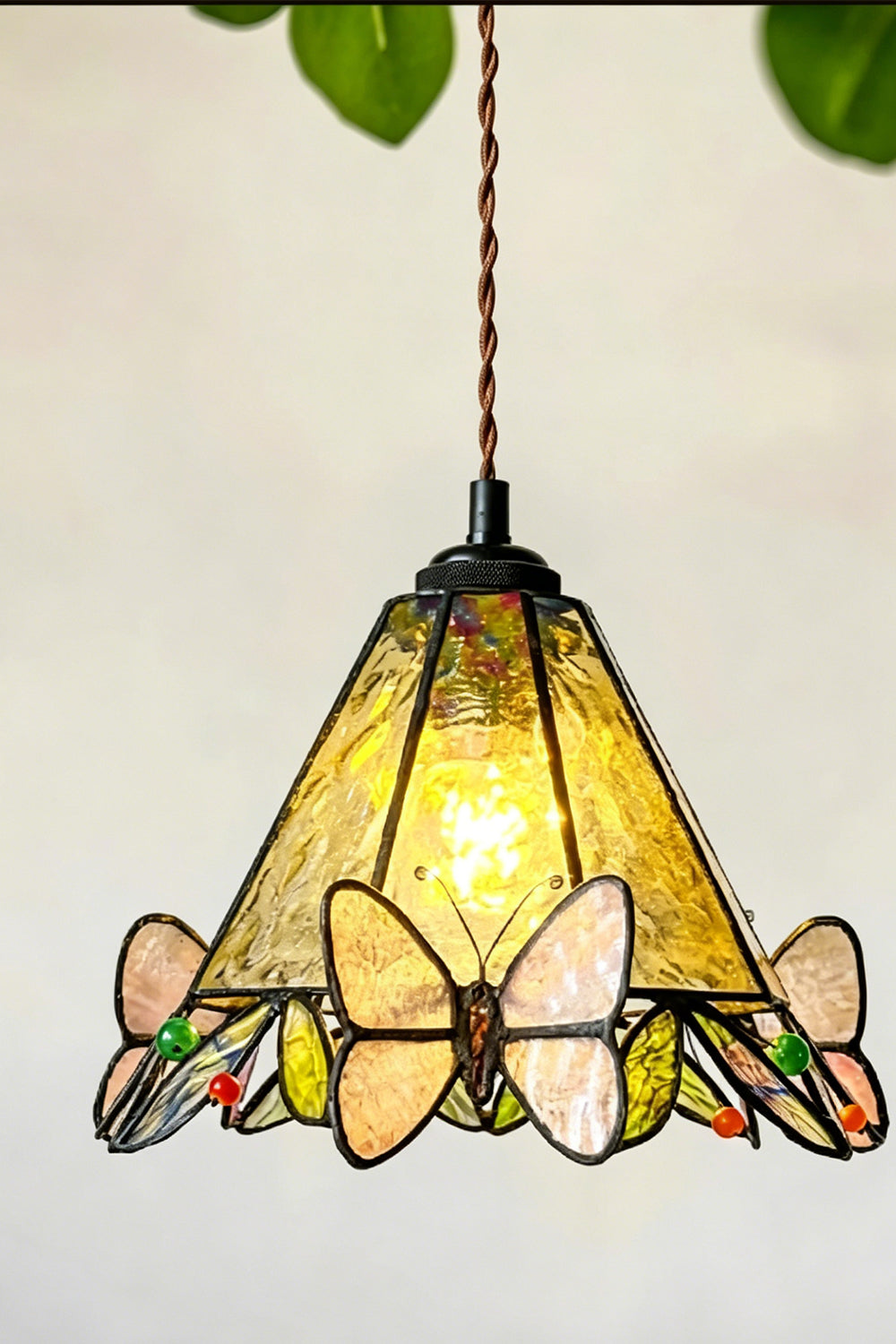Mariposa Canopy Stained Glass Pendant Lamp | Glass, Metal | Closer view of the glowing butterfly pendant lamp hanging with green plant leaves peaking into the frame | Samu Lamp