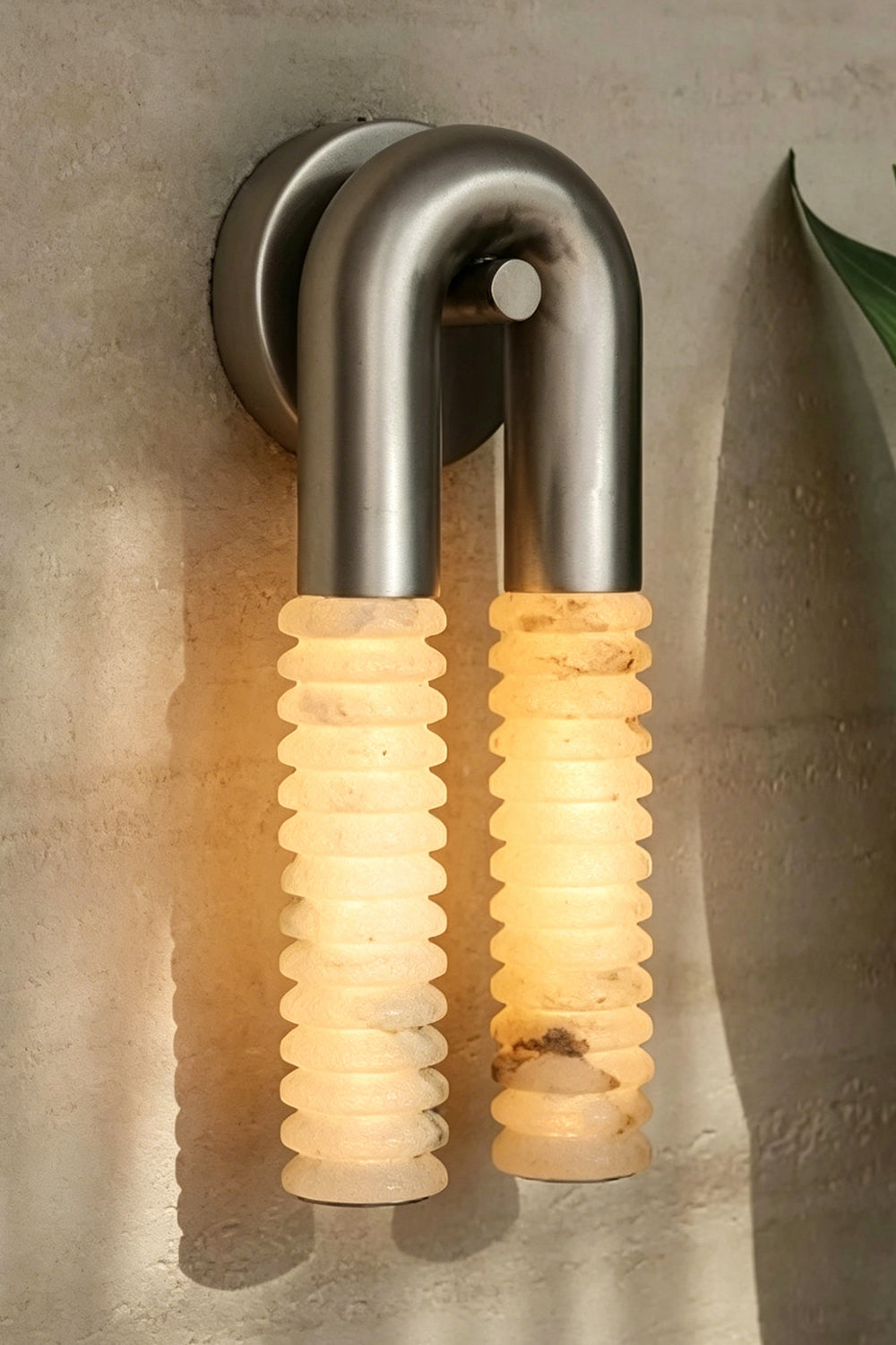 Monolith Arc Alabaster Wall Lamp | Alabaster, Metal | Side profile showing the depth of the metal arc and ribbed stone tubes on a textured wall | Samu Lamp