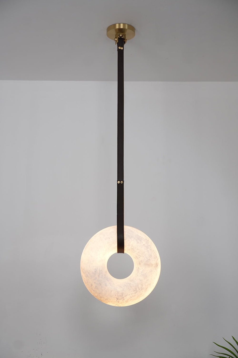 Nerissa Alabaster Ring Leather Pendant Light | Natural Alabaster, Leather and Brass | Single-strap disc variant illuminated softly on a grey wall | Samu Lamp