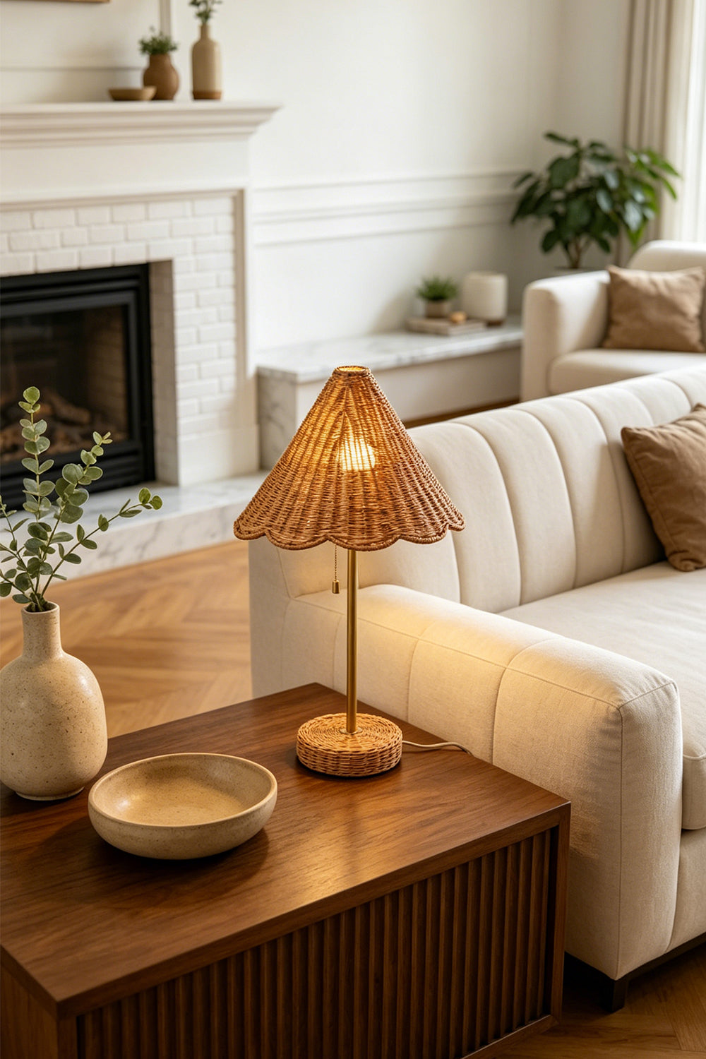 Osrilla Scalloped Rattan Table Lamp | Rattan, Brass | Fully illuminated bohemian rattan table lamp providing warm ambient light on a wooden end table next to a cream sectional sofa near a fireplace | Samu Lamp