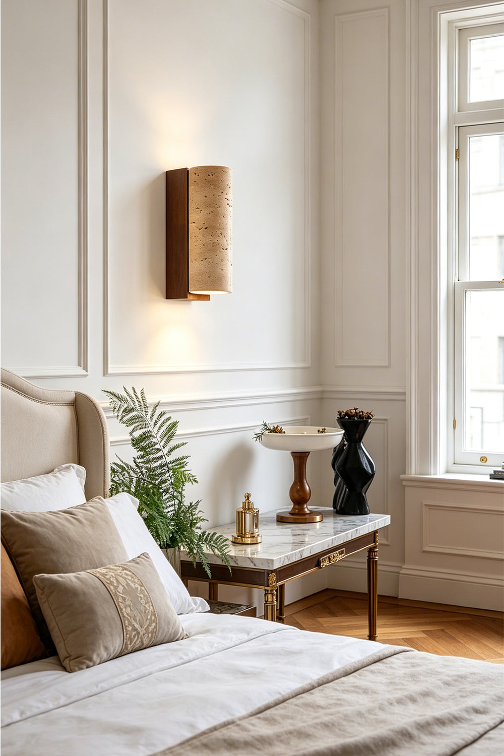 Outcrop Beige Travertine Wall Sconce | Travertine, Metal | Mounted as an elegant bedside accent light casting a warm upward and downward glow on a white wall next to a bed with a cream headboard | Samu Lamp