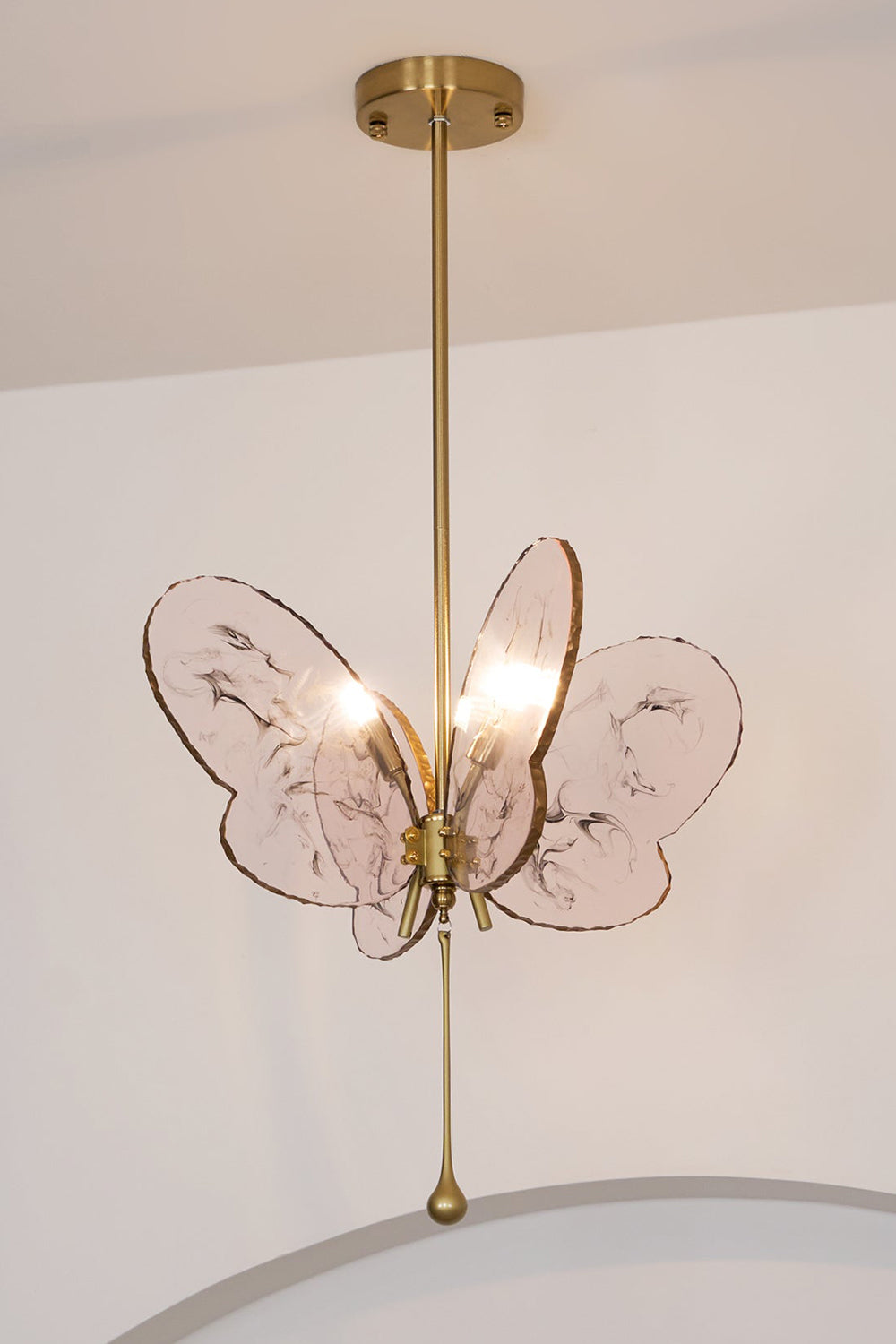 Papillon Glass Butterfly Pendant Lamp | Glass, Metal | Low-angle view looking up at the fully lit pink butterfly hanging light against a plain white wall | Samu Lamp