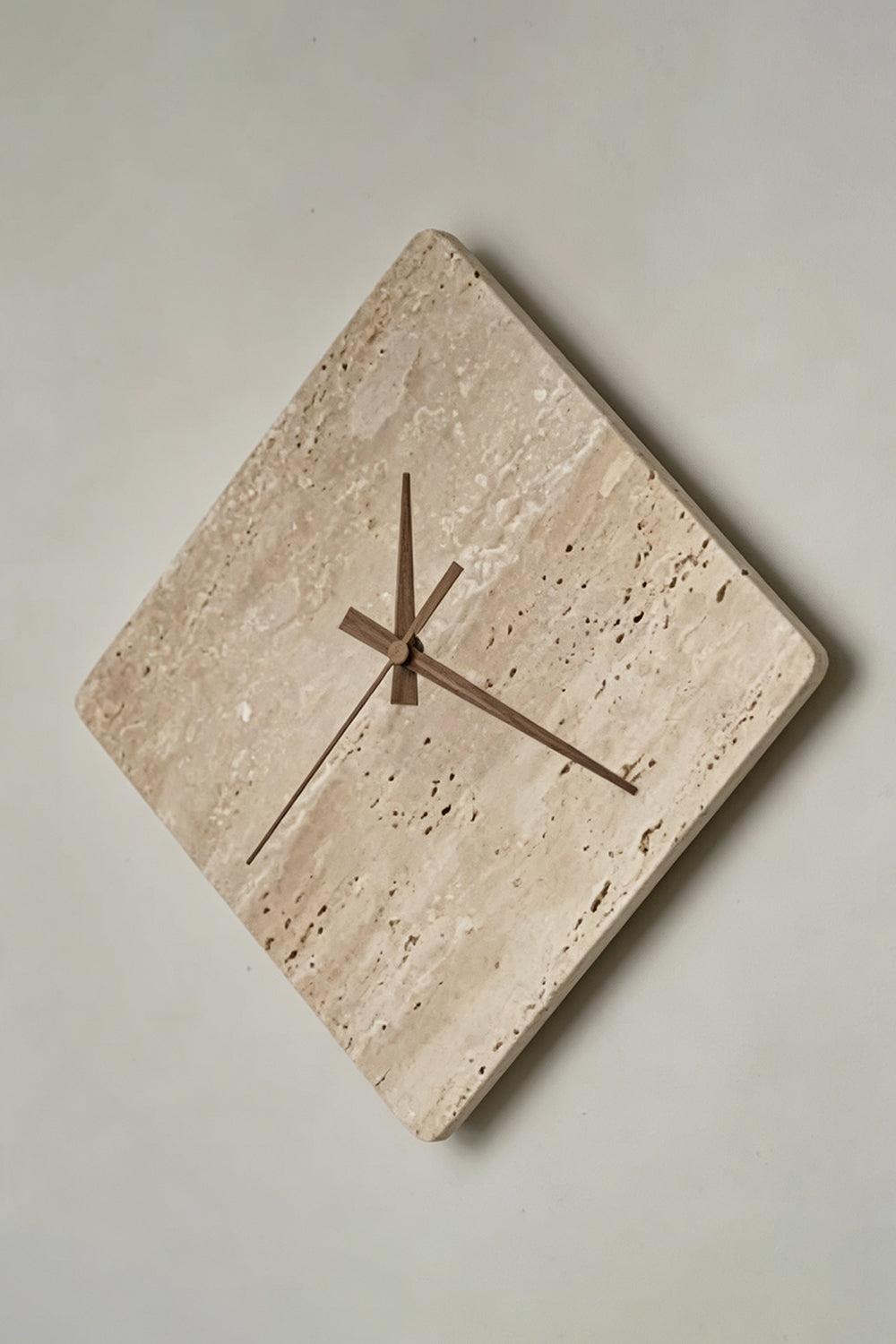 Petra Compass Travertine Backlit Wall Lamp | Travertine, Iron | Angled frontal view of the unlit travertine clock-lamp displaying its natural porous stone surface against a grey wall | Detail shot | Samu Lamp