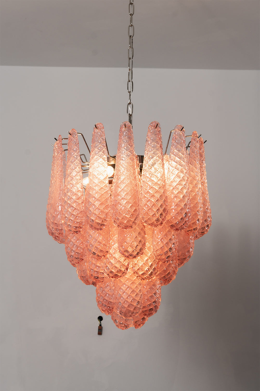Amara Blush Cascading Glass Chandelier | Glass, Metal | Medium shot of the lit tiered chandelier radiating a soft rose glow against a light grey background | Samu Lamp