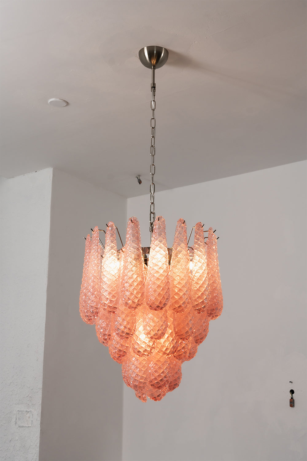 Amara Blush Cascading Glass Chandelier | Glass, Metal | Close-up frontal view of the glowing blush pink cascading chandelier against a plain grey wall | Samu Lamp