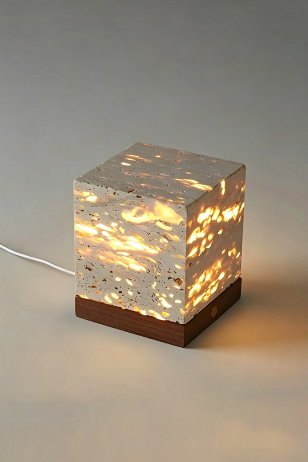Radiant Monolith Travertine Table Lamp | Travertine, Wood | Lit front view on plain background | Intricate internal light pattern | Dark wood base, gold button | Samu Lamp