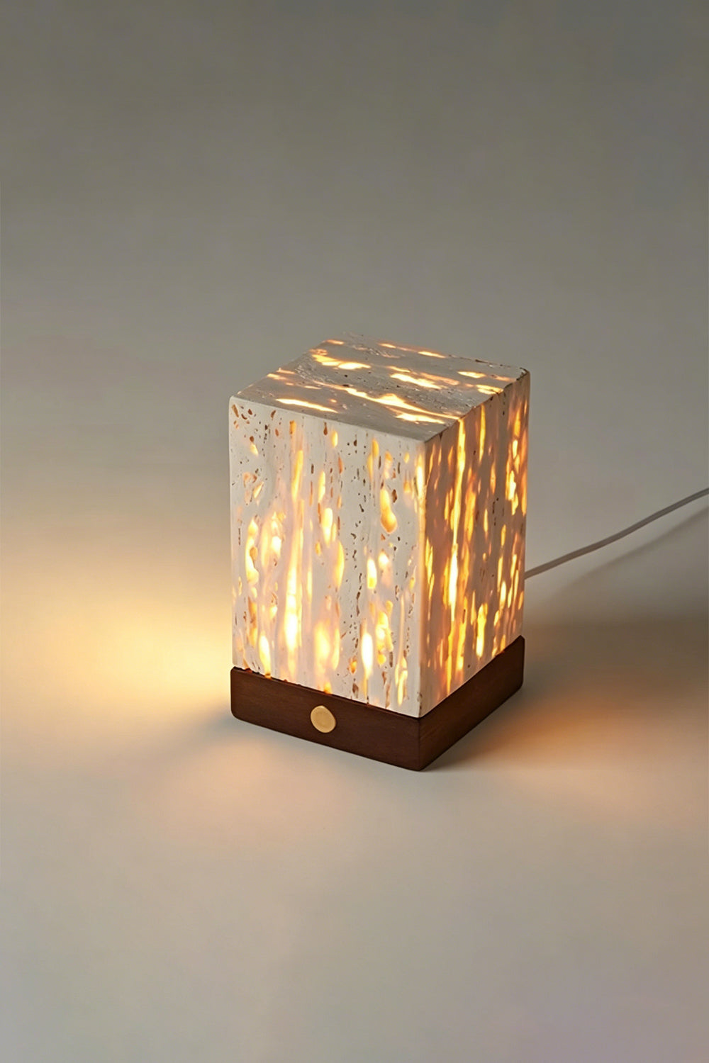 Radiant Monolith Travertine Table Lamp | Travertine, Wood | Lit angled view on grey surface | Intricate internal light pattern | Dark wood base, warm glow | Samu Lamp
