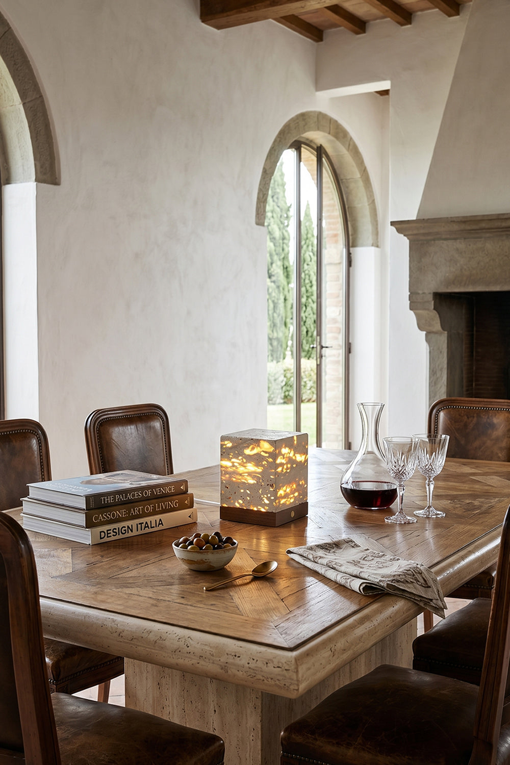 Radiant Monolith Travertine Table Lamp | Travertine, Wood | Lit on travertine dining table | Tuscan-style dining interior | Rustic stone walls, hill view | Samu Lamp