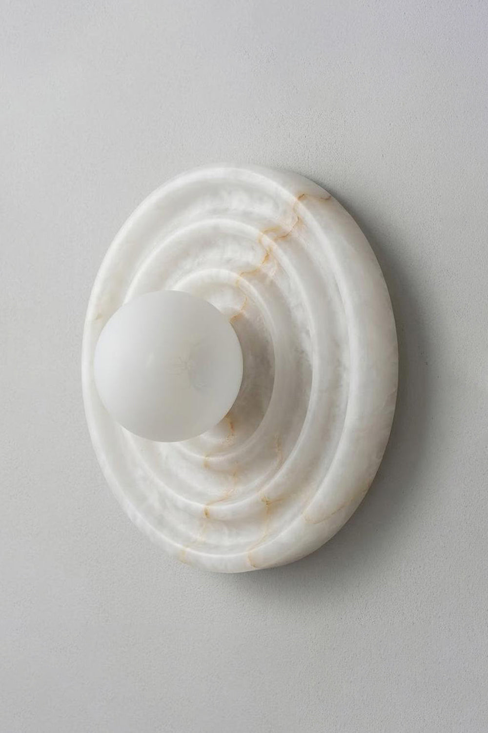 Rhea Rippled Alabaster Wall Sconce | Studio Profile View | Side angle view of the unlit minimalist circular stone wall fixture flush mounted against a clean light grey backdrop wall | Samu Lamp