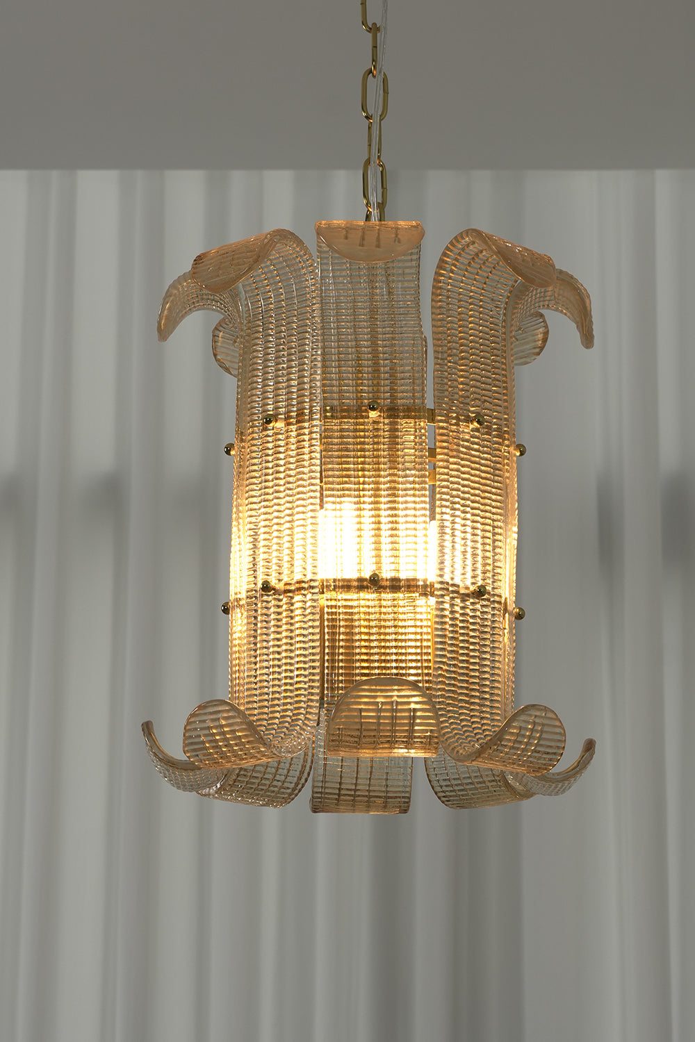 Lit ribbed glass scroll chandelier against white curtain | Metal | Glass | Front view showing the elegant scroll shape and warm glow | Samu Lamp