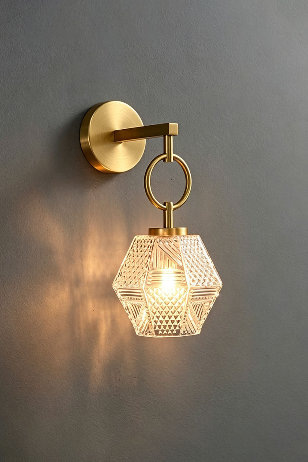 Rosamund Faceted Glass Wall Sconce | Faceted Glass and Brass | Brass finish sconce glowing warmly against a dark grey backdrop | Samu Lamp