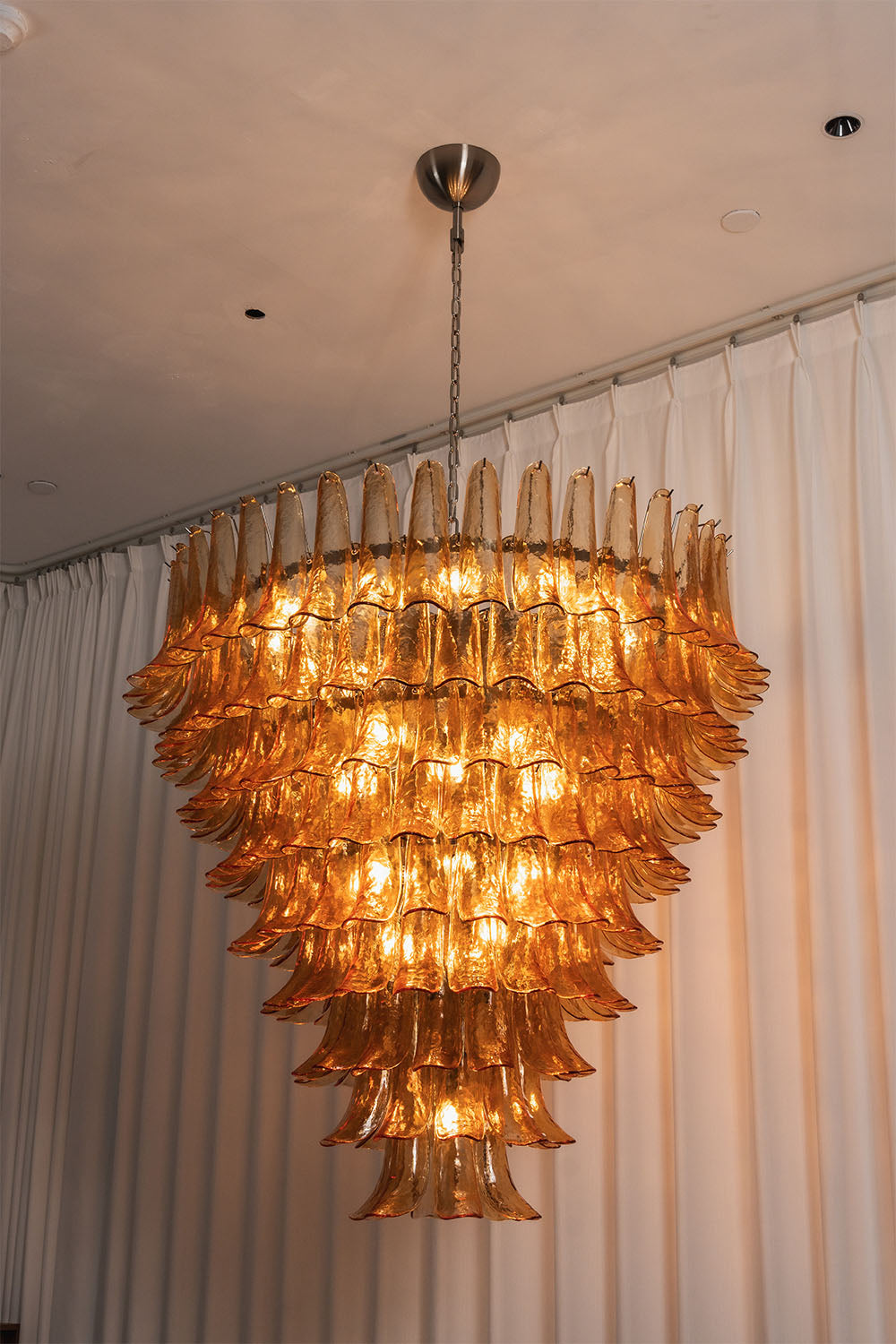 Rouveline Rippled Amber Glass Cone Chandelier | Rippled Amber Glass and Metal | Real-life view glowing warmly next to a sheer white curtain | Samu Lamp