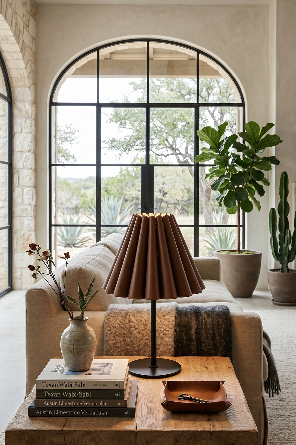 Rowan Leather Pleated Table Lamp | Leather, Metal | Positioned on a wooden coffee table in a modern lounge space featuring a large black framed arched window | Samu Lamp