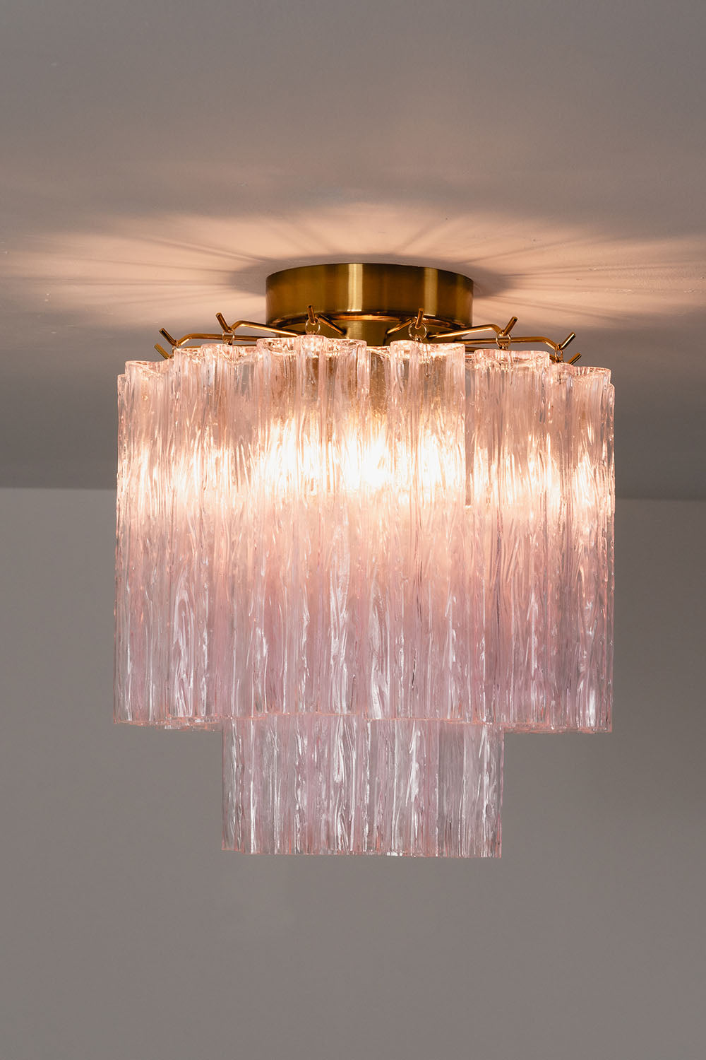 Seraphine Starburst Glass Tube Ceiling Light | Pink Glass and Brass | Studio display showing the chandelier glowing warmly against a dark grey wall | Samu Lamp