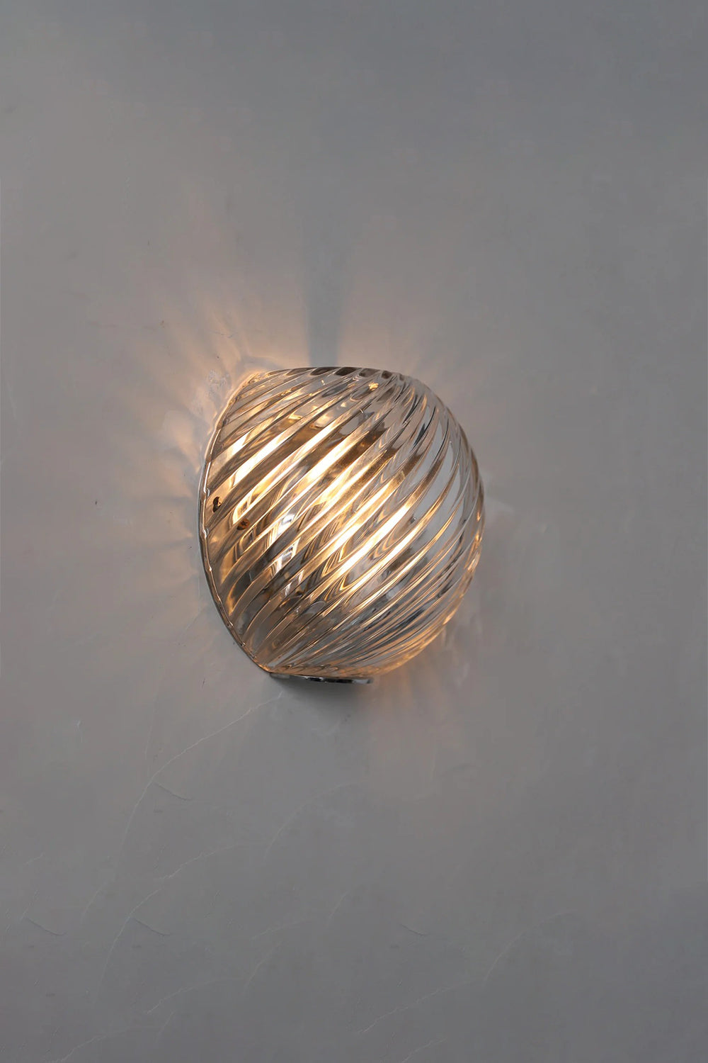 Silas Ribbed Glass Wall Sconce | Ribbed Glass, Metal | Staged lit studio view of a clear glass variant casting beautiful swirled ribbed light patterns against a grey wall | Samu Lamp