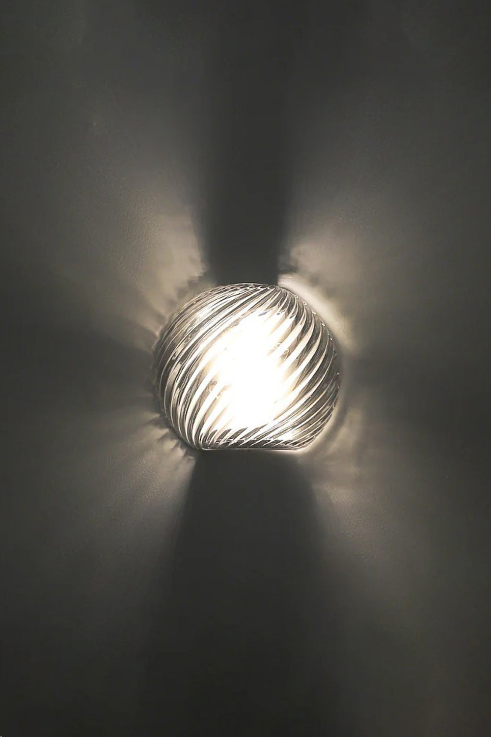 Silas Ribbed Glass Wall Sconce | Ribbed Glass, Metal | Close-up illuminated view against a dark charcoal backdrop, showing the dramatic starburst ribbed light patterns cast on the wall | Samu Lamp
