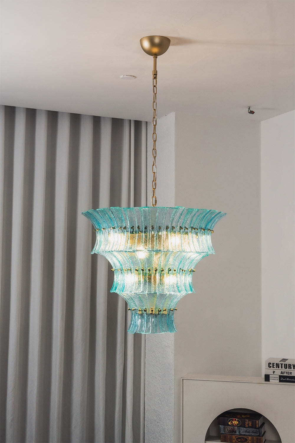 Thalassa Aquamarine Textured Glass Chandelier | Glass, Metal | Alternative studio shot of the illuminated tiered architectural glass light fixture hanging elegantly next to a grey drapery panel | Samu Lamp