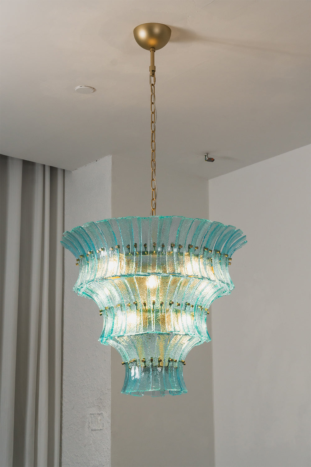 Thalassa Aquamarine Textured Glass Chandelier | Glass, Metal | Alternative studio shot of the illuminated tiered architectural glass light fixture hanging elegantly next to a grey drapery panel | Samu Lamp