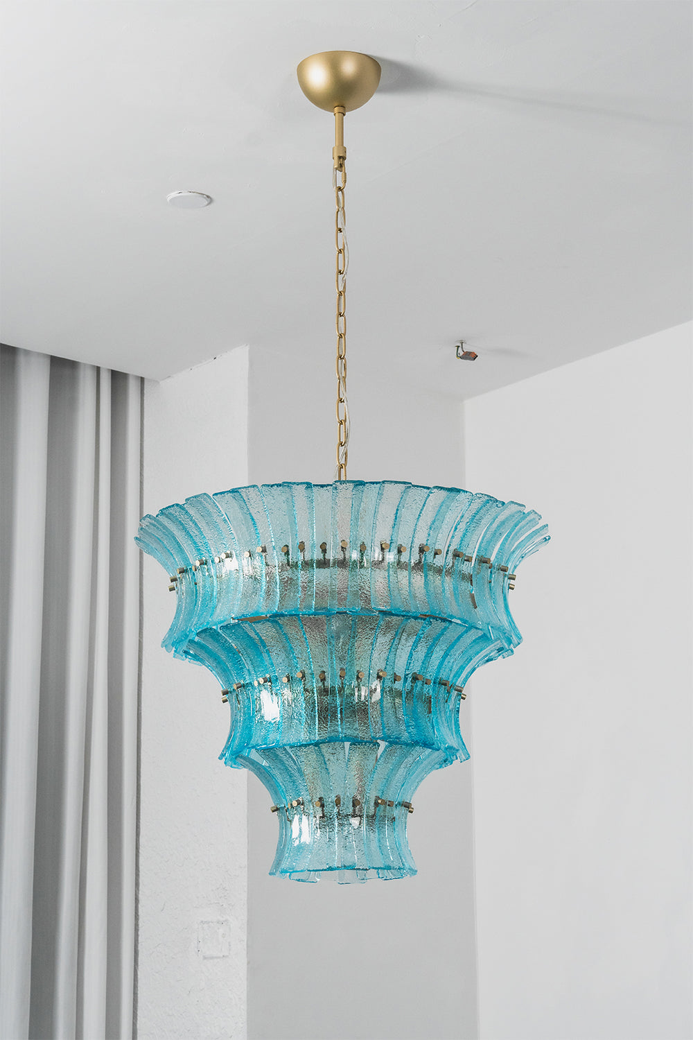 Thalassa Aquamarine Textured Glass Chandelier | Glass, Metal | Studio staged view of the tall multi-tiered lit aquamarine glass chandelier casting a rich sea-colored glow near grey window curtains | Samu Lamp