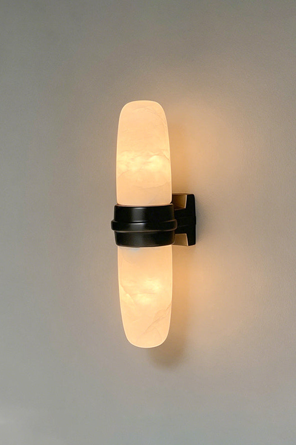 Theron Linear Alabaster Wall Lamp | Alabaster, Metal | Vertical shot of the illuminated alabaster sconce with a dark black center band on a plain grey backdrop | Samu Lamp