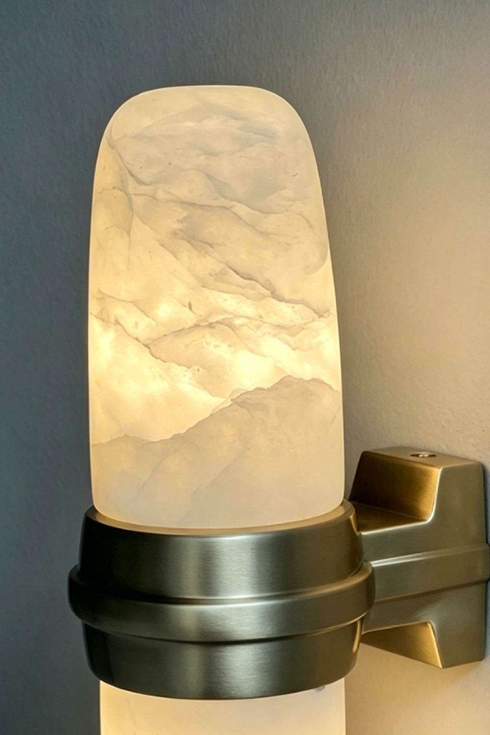 Theron Linear Alabaster Wall Lamp | Alabaster, Metal | Moody close-up focus on the dark bronze mounting band holding the glowing translucent stone capsule under low light | Detail shot | Samu Lamp