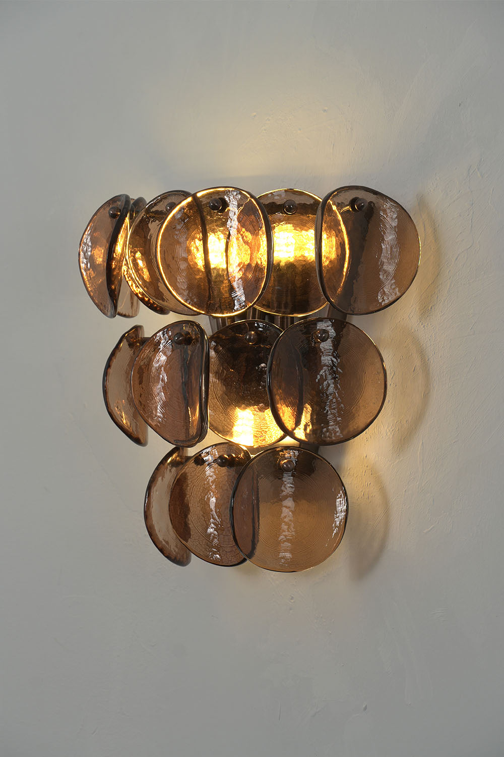 Tisanne Tiered Glass Disc Wall Lamp | Smoked Glass and Metal | Real-life installation view glowing softly against a light grey wall | Samu Lamp
