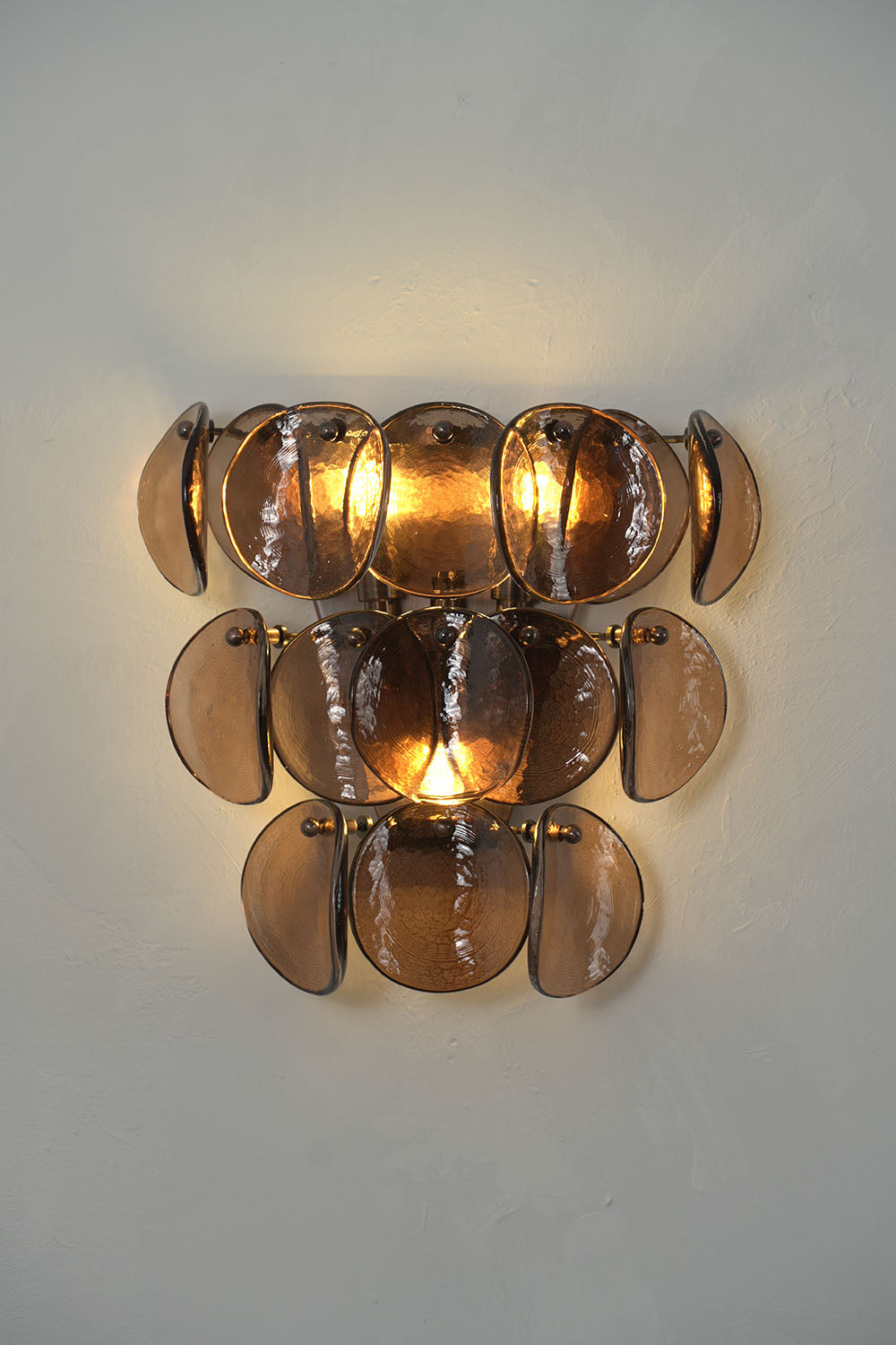 Tisanne Tiered Glass Disc Wall Lamp | Smoked Glass and Metal | Front view illuminated warmly on a plain neutral wall backdrop | Samu Lamp