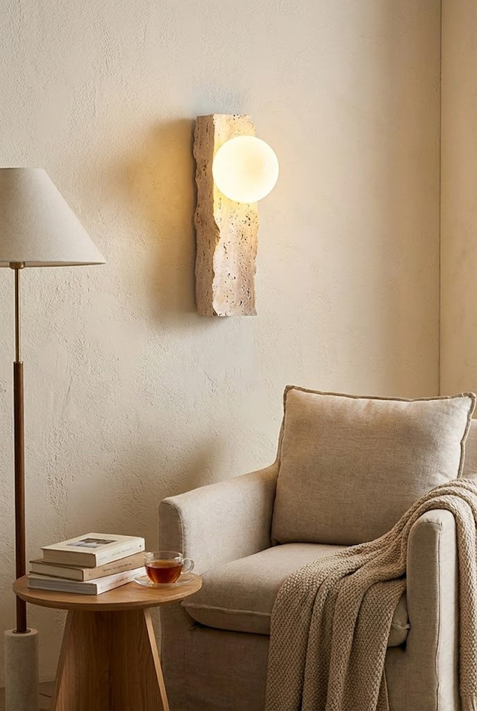 Lit travertine block sconce in a reading nook | Yellow Travertine, Metal, Glass | Mounted above a cozy armchair and floor lamp | Samu Lamp